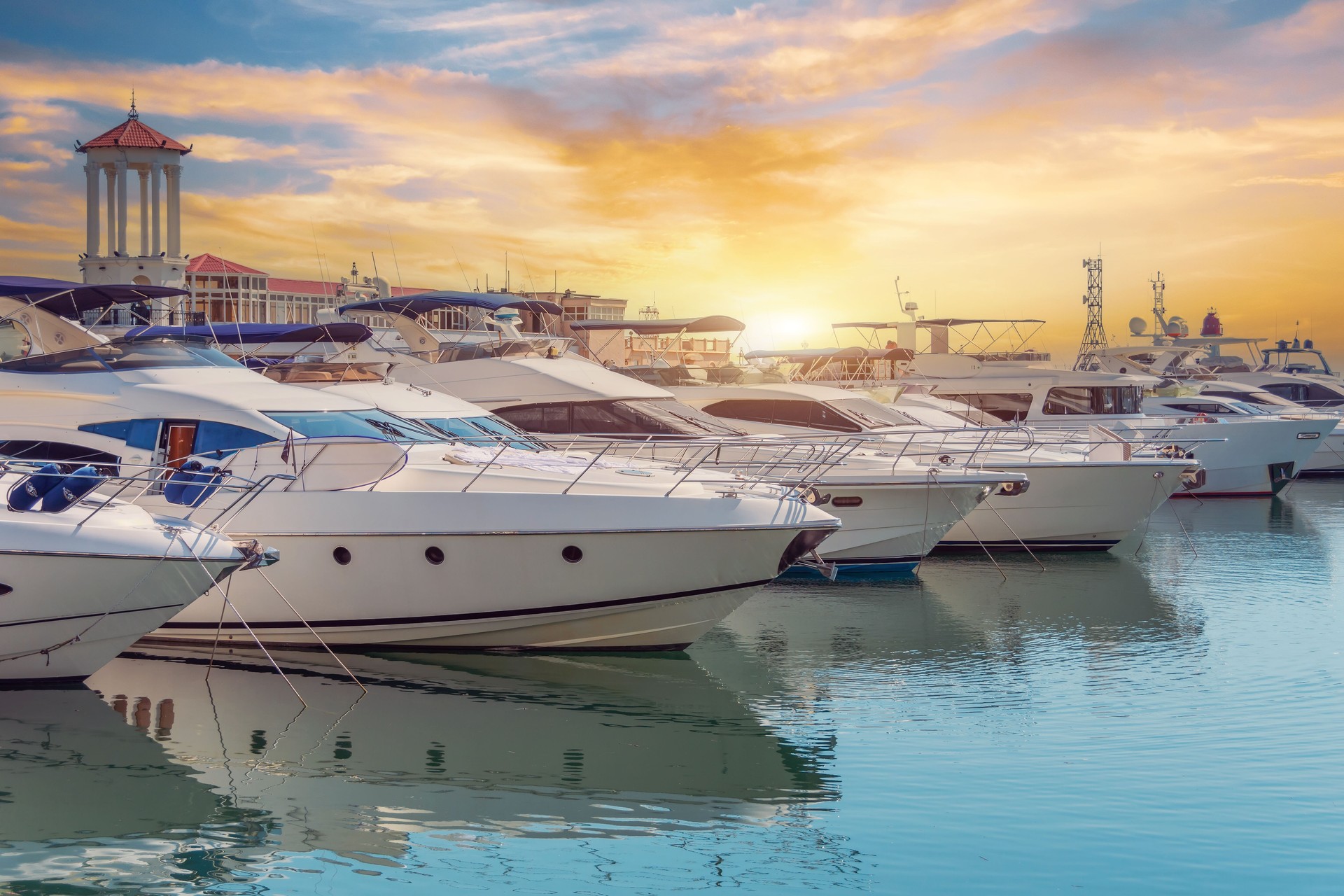 The blue water reflects orange sunset sky. Rows of sailing yachts, motor boats and fishing boats. The water area of port